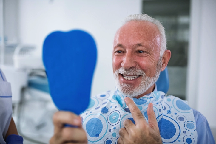 senior dental patient smiling and pointing at teeth while holding a mirror