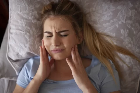 young woman laying in bed and holding jaw with both hands